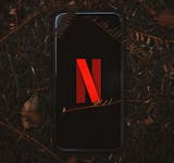 Smartphone with Netflix logo on screen, surrounded by dry leaves outdoors.